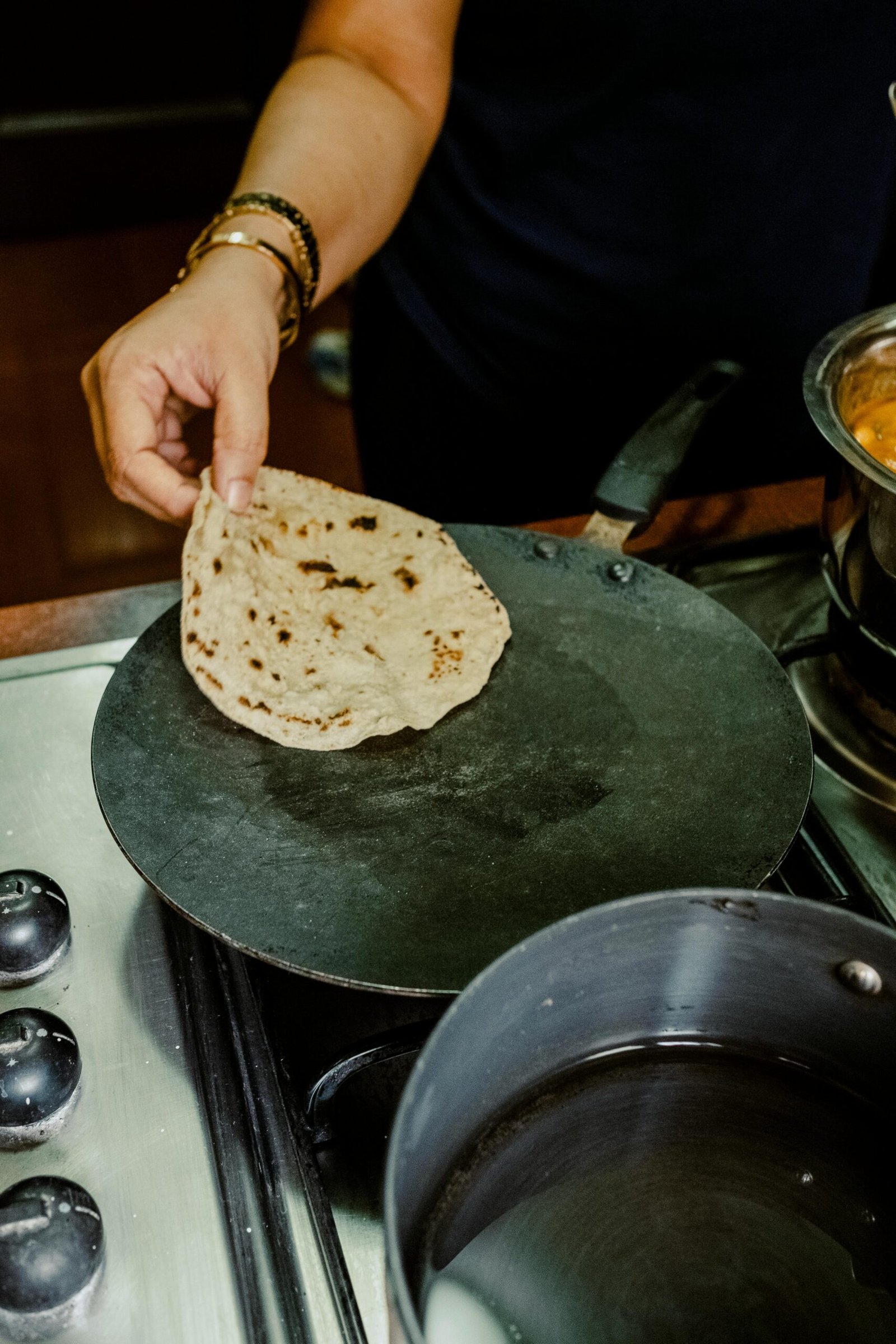 Indian woman with gold bangles making roti on a tawa with dal in background for family meal